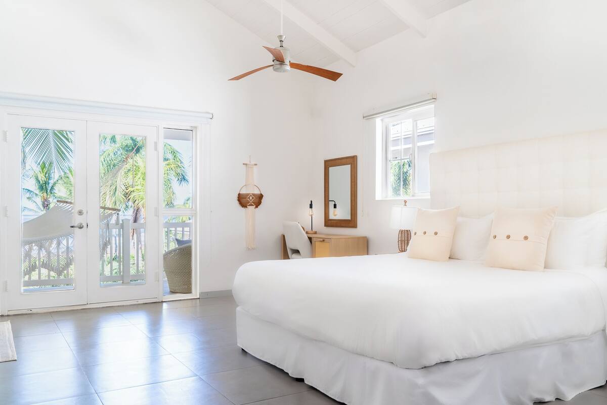 A spacious bedroom with a large bed, a desk, and a chair. There is a ceiling fan, a window, and a door leading to a balcony.