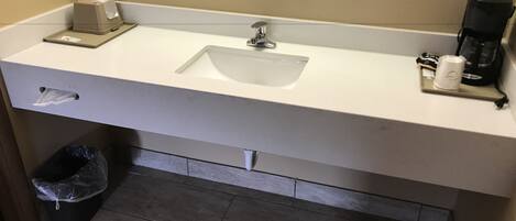 A bathroom with a white sink, a mirror, a trash bin, and a coffee maker.