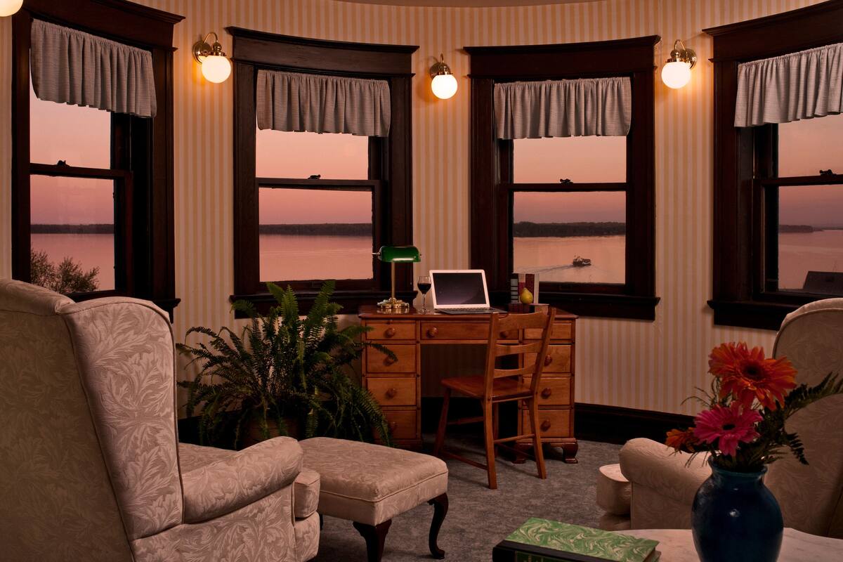 A room with a desk, two chairs, a laptop, a vase with flowers, and a view of a body of water through windows.
