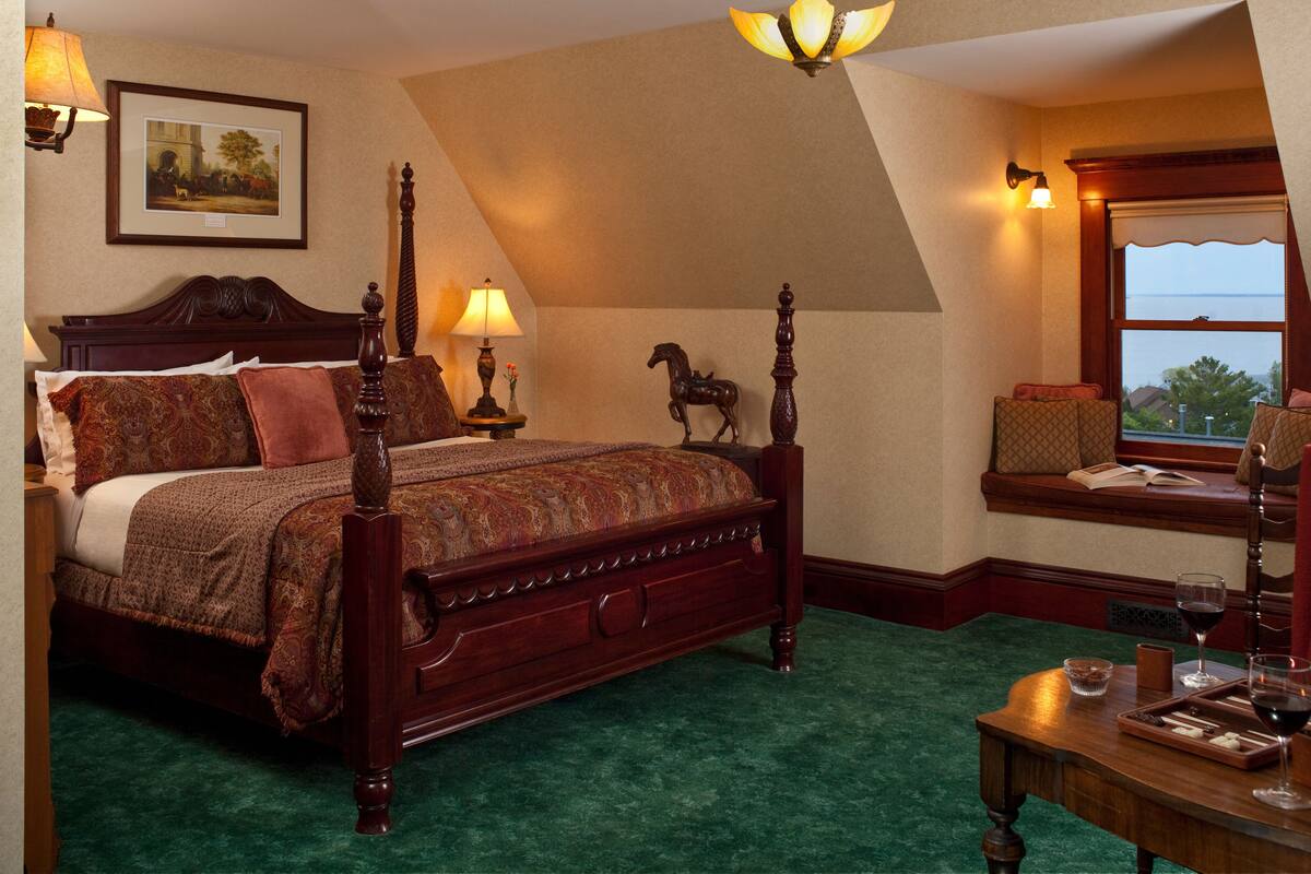 A bedroom with a large bed, a window seat with a view, and a small table with a glass of wine.