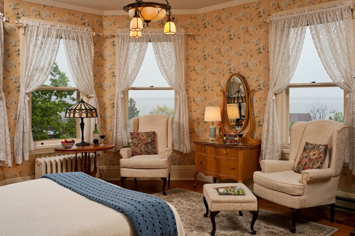 A bedroom with a large bed, two armchairs, a dresser, a mirror, and a view of the outdoors.
