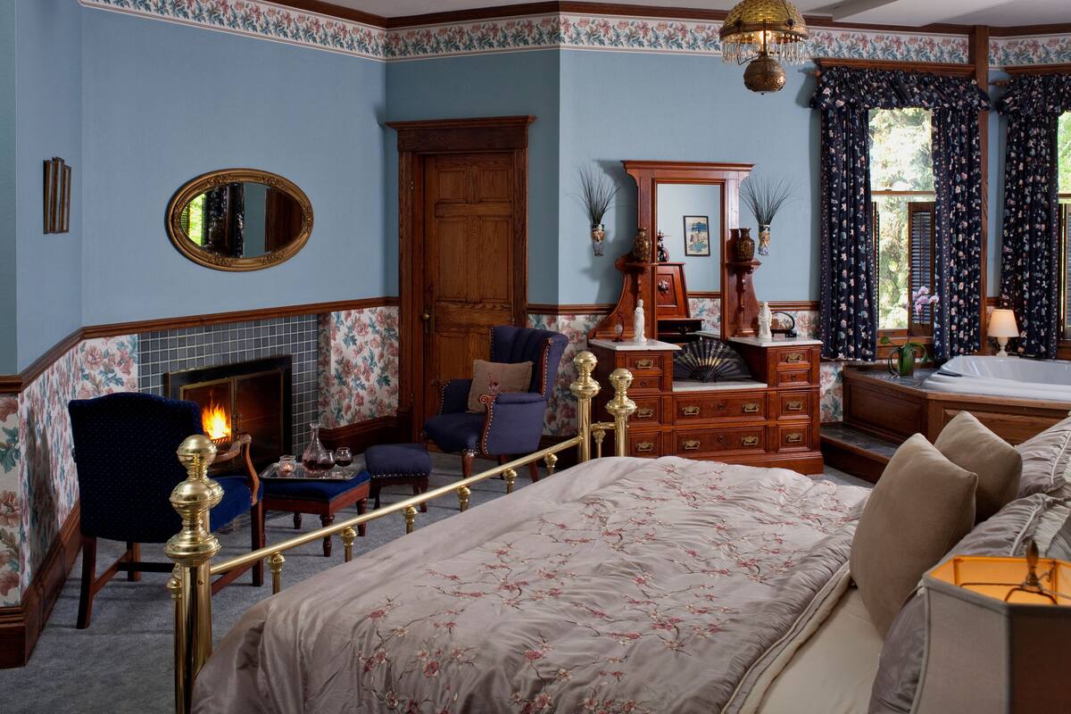 A bedroom with a bed, a chair, a dresser, a mirror, and a fireplace.