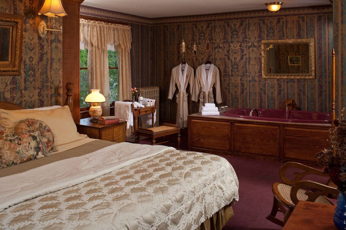 A bedroom with a large bed, a wooden dresser, a vanity with a mirror, and a bathtub.