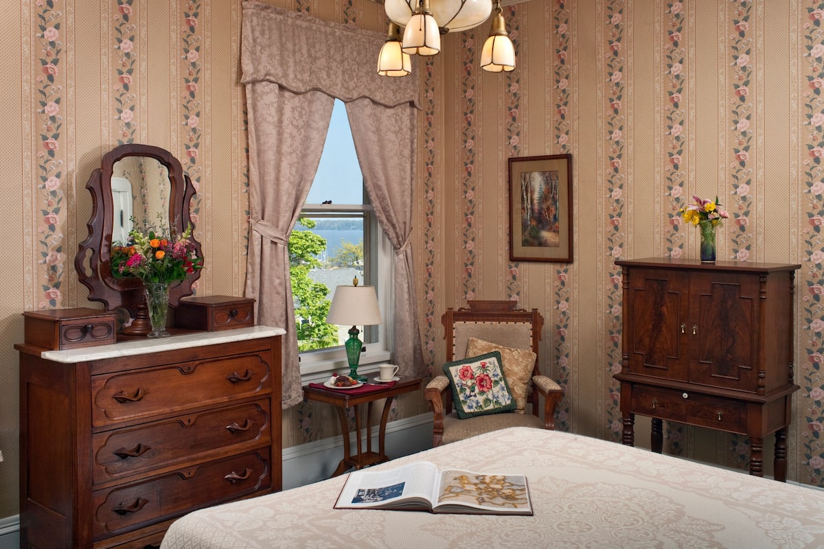 A bedroom with a bed, dresser, vanity, and a window with curtains.
