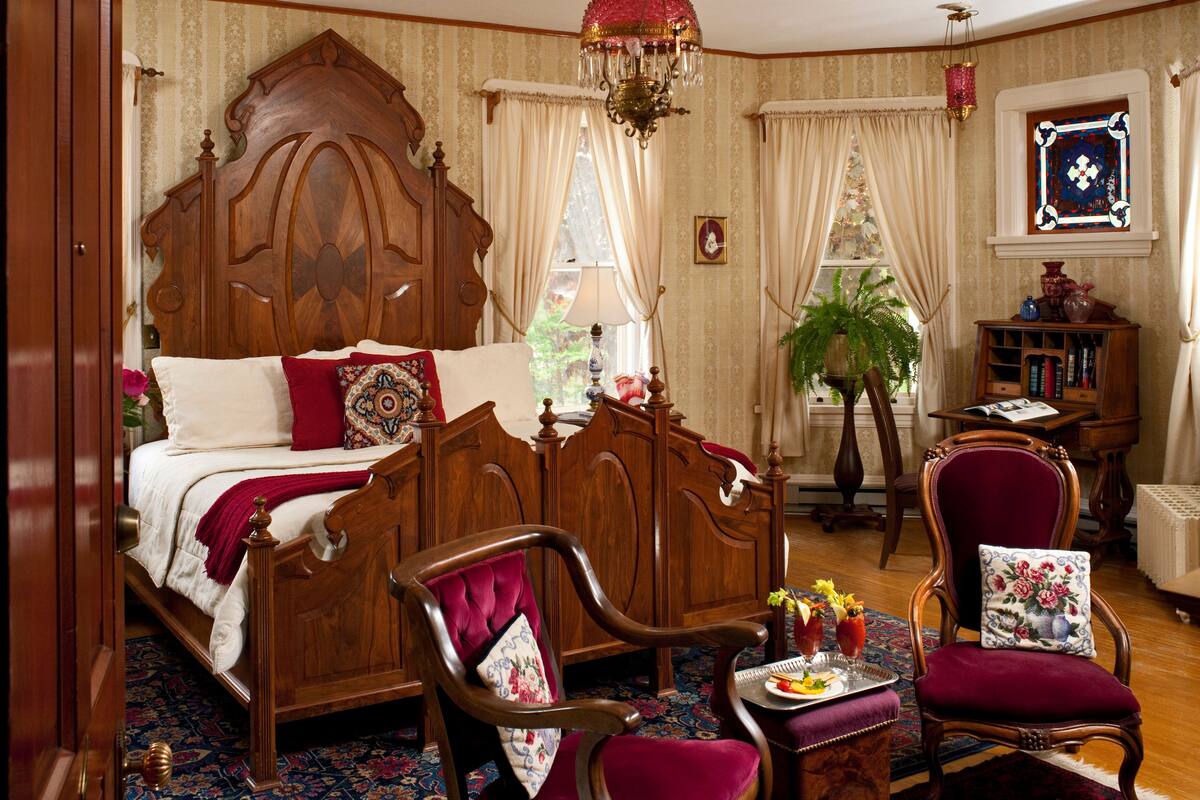 A traditional bedroom with a large wooden bed, two chairs, a small table with a tray, and a potted plant.