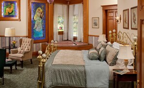 A spacious bedroom with a large bed, a sitting area with a chair, a small table, and a view of the garden through the window.