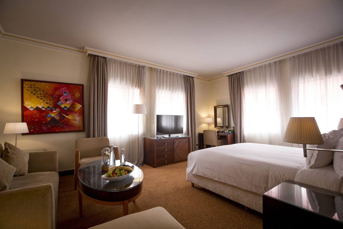 Habitación de hotel con cama, escritorio, televisor, lámpara, silla y un cuadro en la pared.