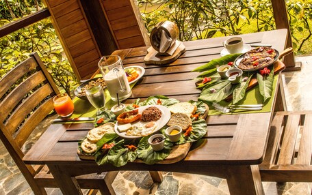 A wooden table set with breakfast, including eggs, toast, and a fruit salad, with a view of greenery in the background.