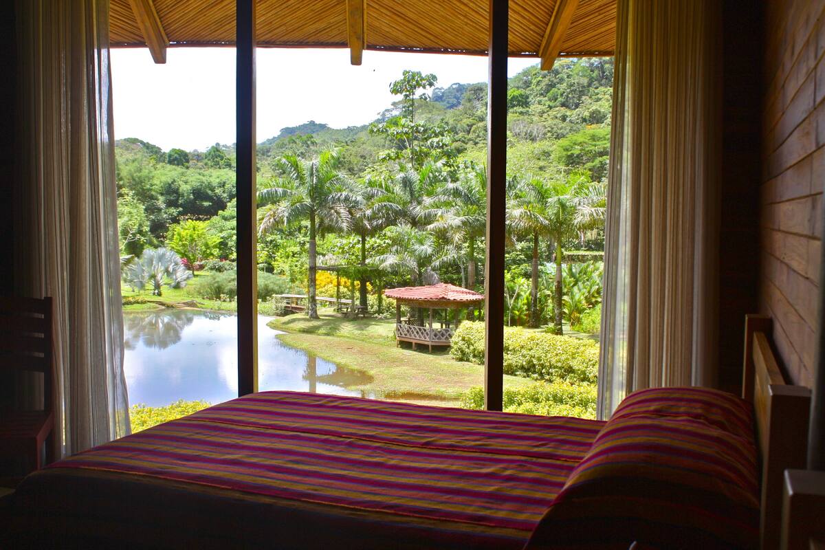 A room with a bed and a view of a lush garden and a pond.