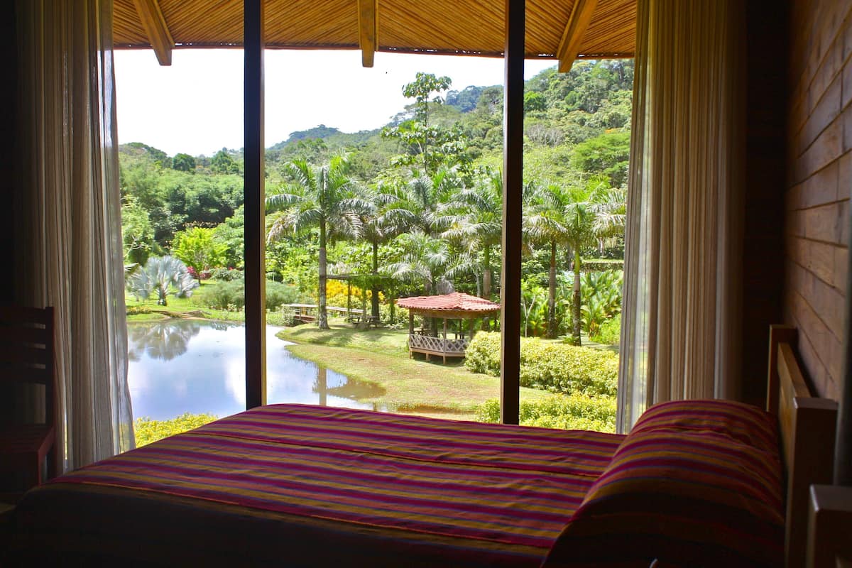 A room with a bed, large windows, and a view of a garden and water body.