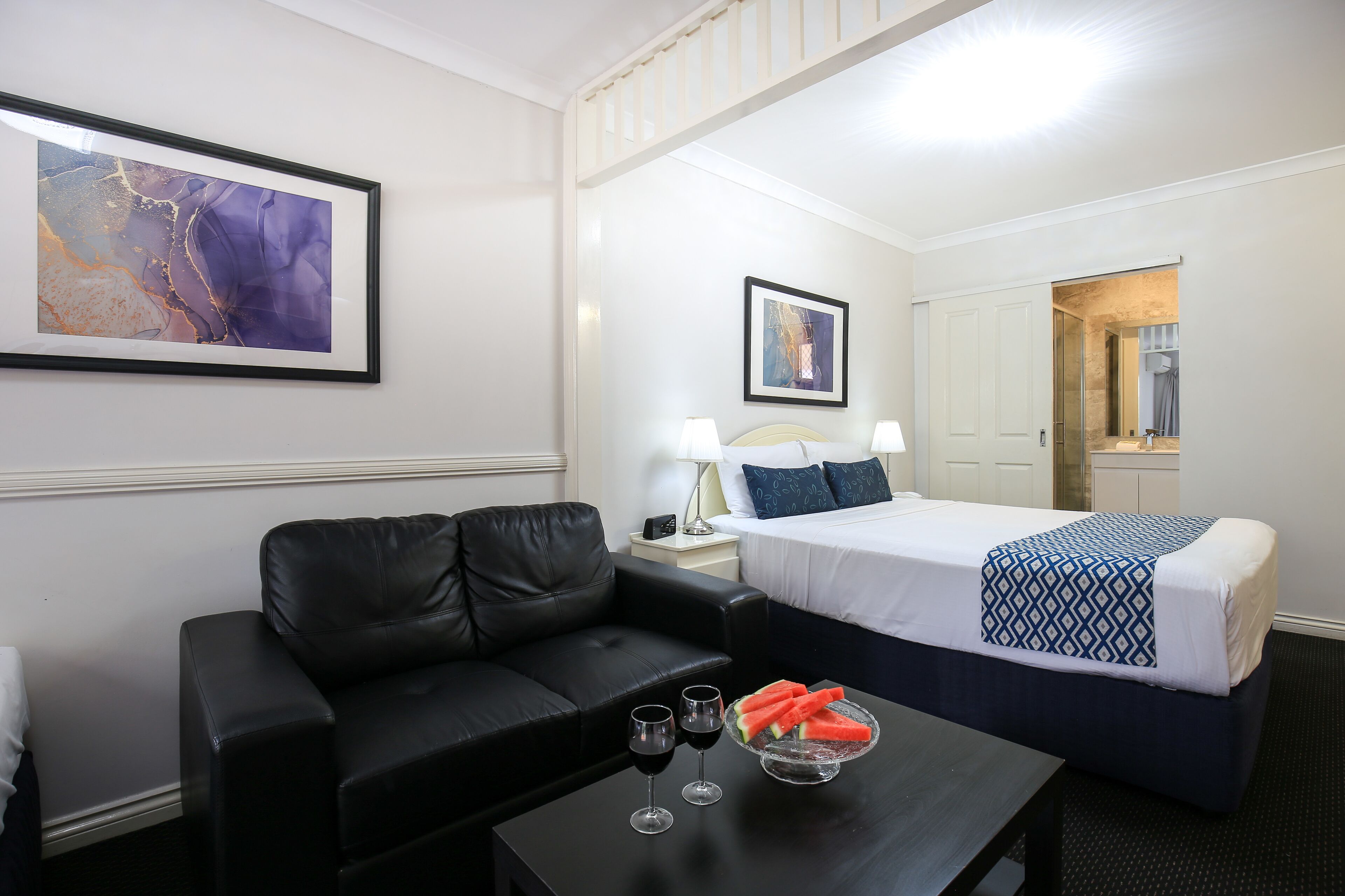 Une chambre d’hôtel avec un canapé en cuir noir, un lit recouvert d’une couverture à motifs, une table de chevet avec une lampe et un tableau abstrait encadré accroché au mur.