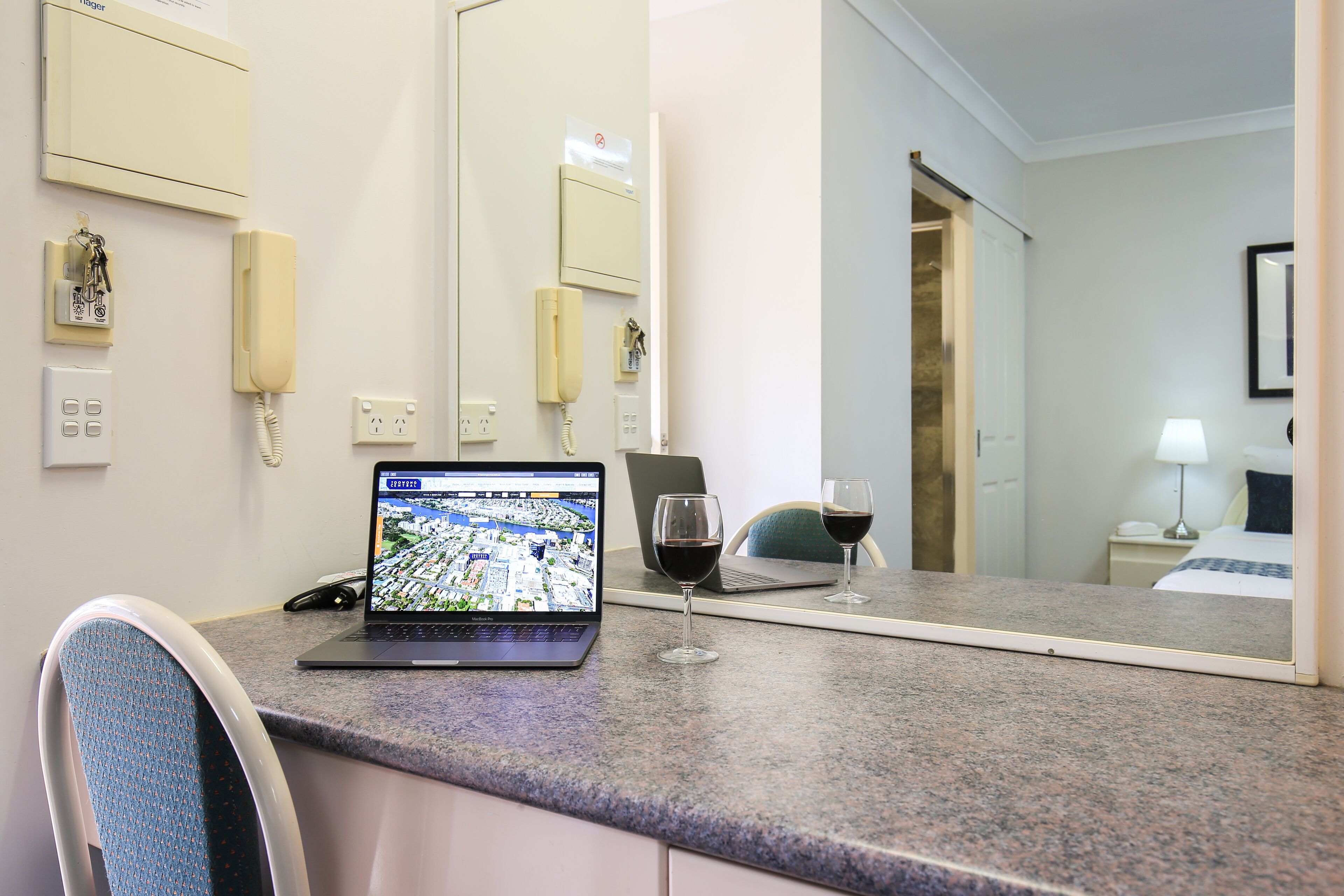 A hotel room with a laptop displaying a map, a wine glass, a phone, and a mirror reflecting the room.
