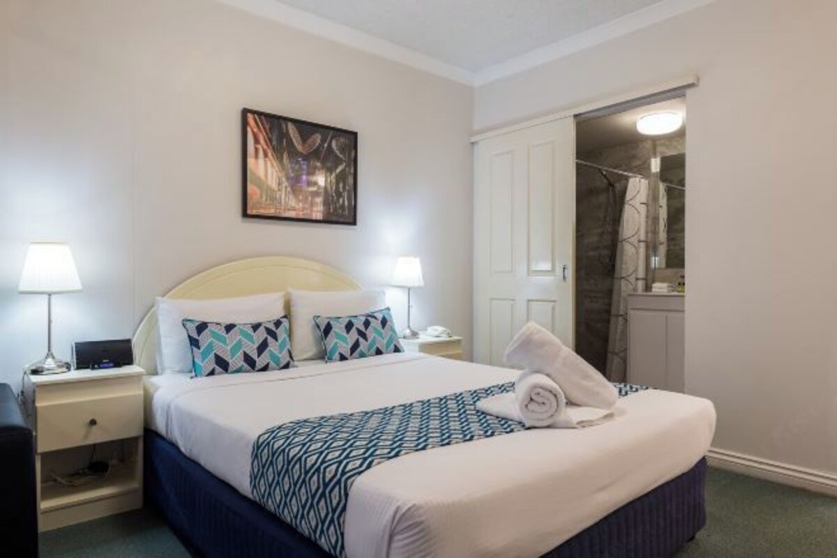 A neatly arranged hotel room with a bed, bedside lamps, a painting on the wall, and a bathroom visible through an open door.