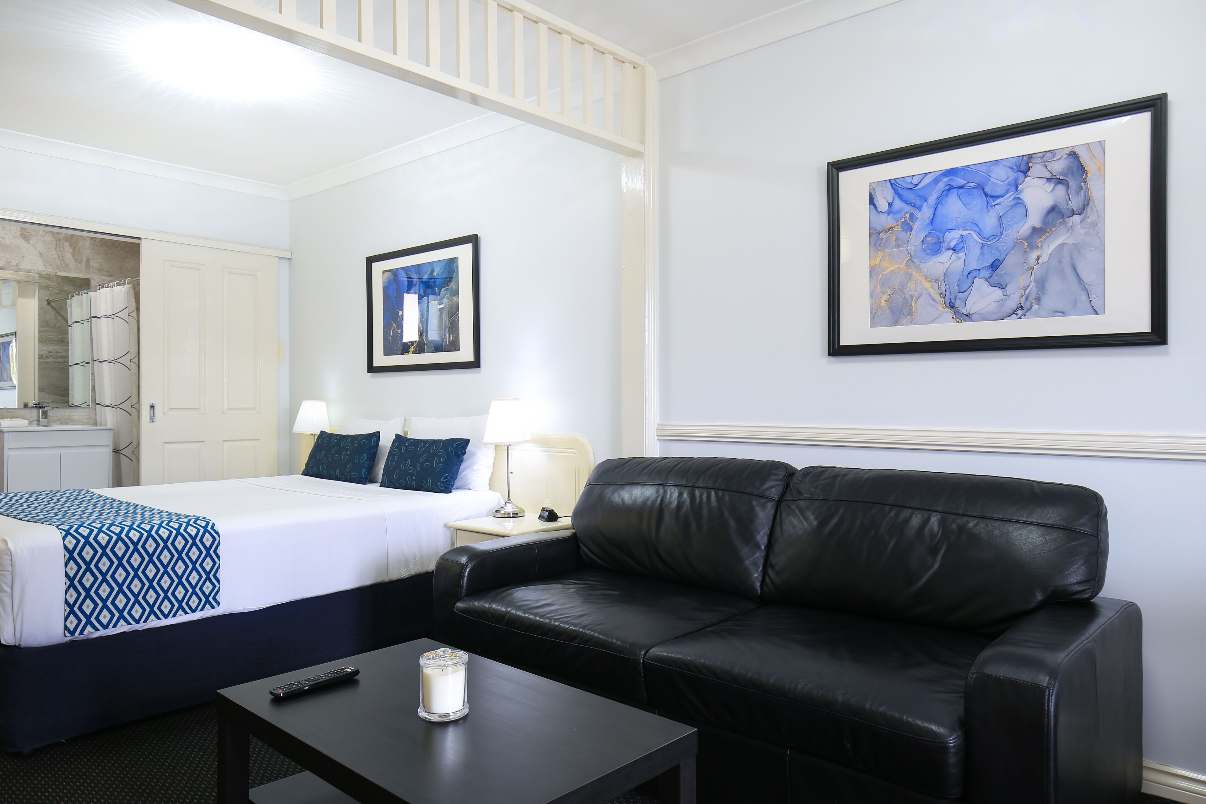 Una habitación de hotel con una cama, un sofá de cuero negro, una mesita de noche con un control remoto y una vela, y dos obras de arte enmarcadas en la pared.