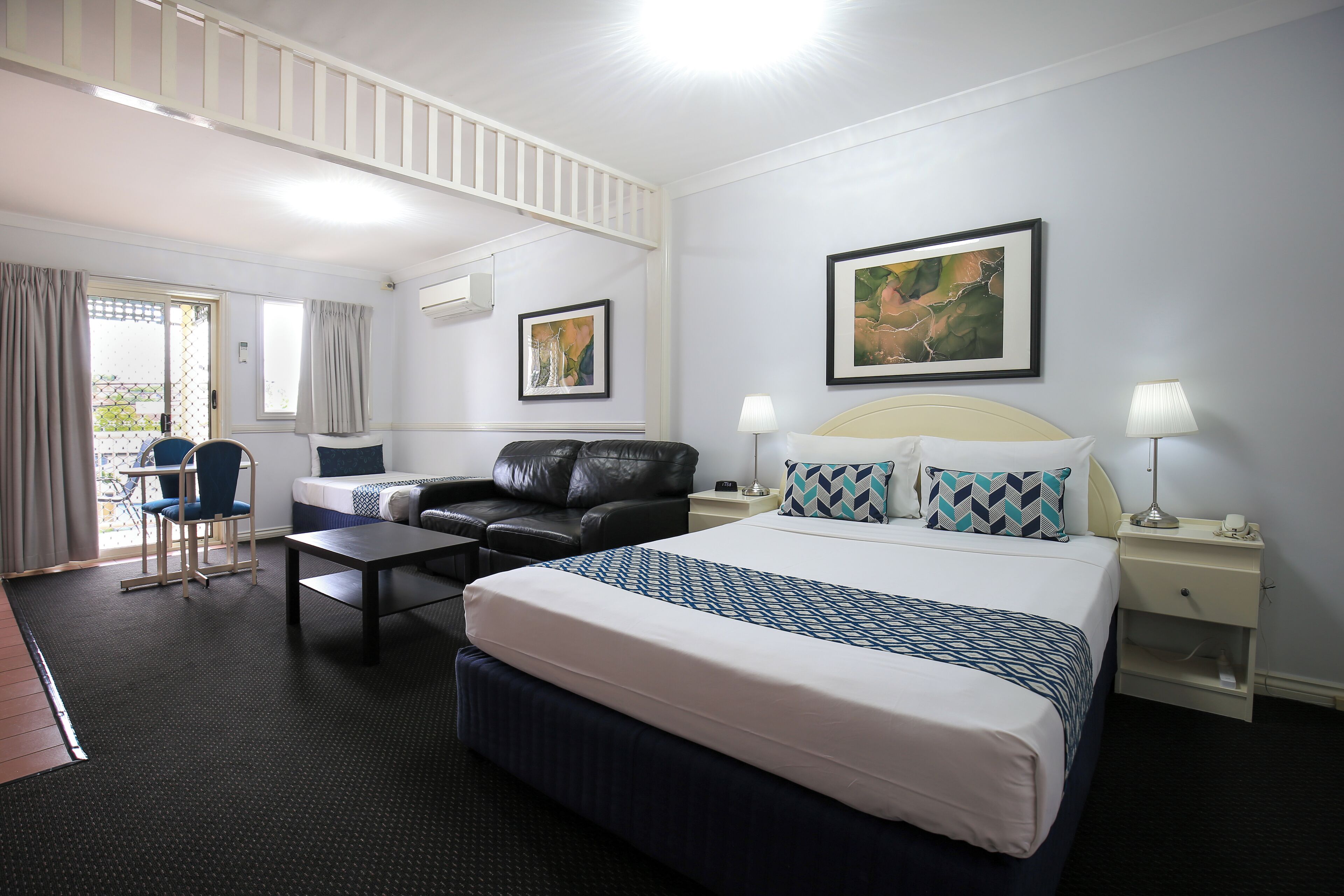 A hotel room with a large bed, a sofa, a desk, and a chair. There are two paintings on the walls, a window with curtains, and a nightstand with a lamp.