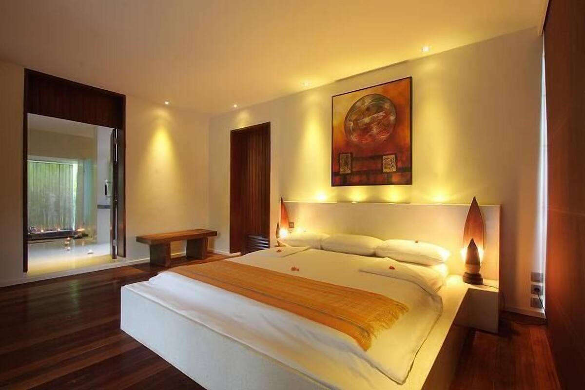Une chambre à coucher moderne avec un grand lit, une table de chevet, un banc et une œuvre d’art accrochée au mur.