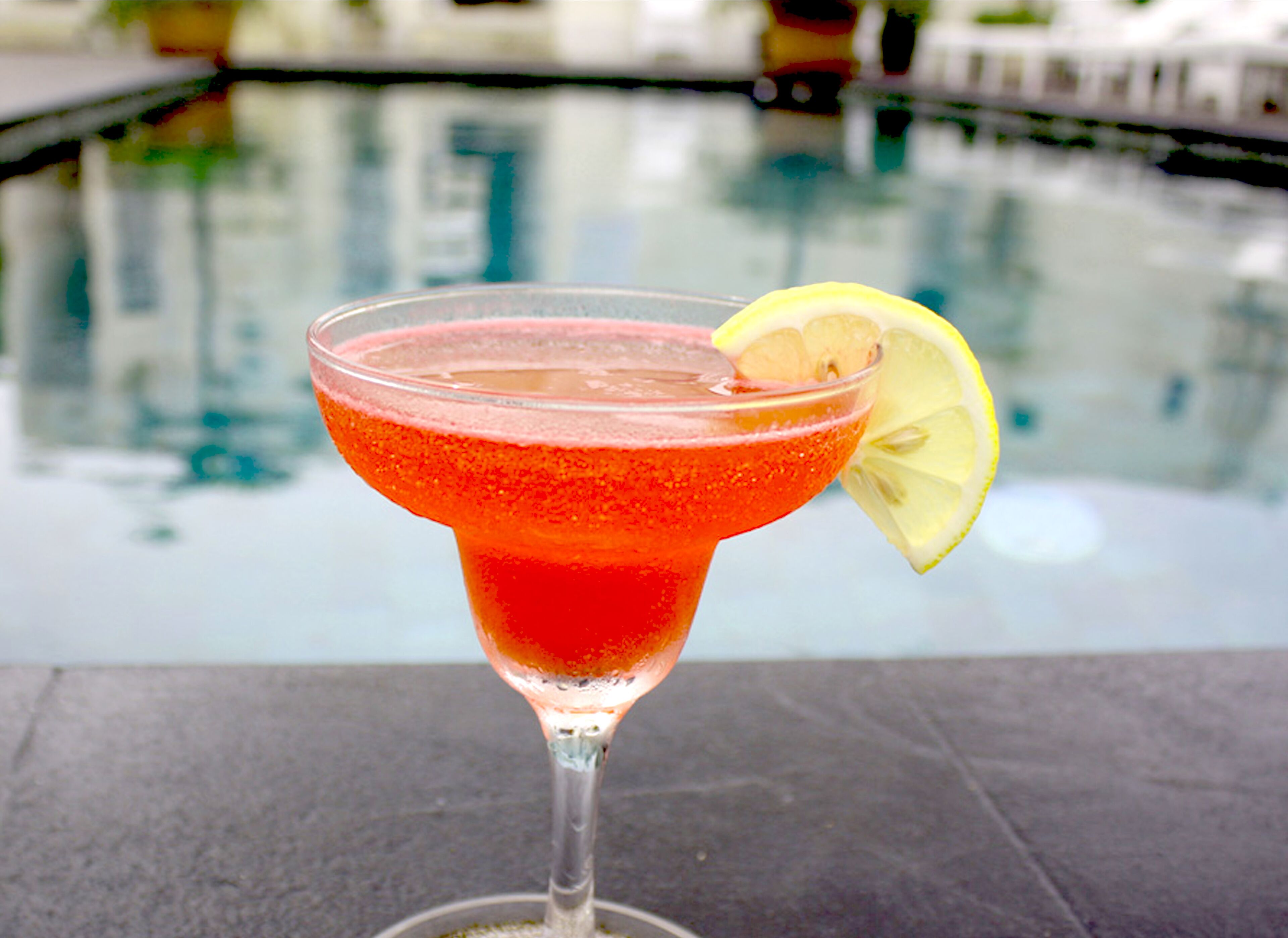 A cocktail glass with a slice of lemon on the rim, placed on a stone surface near a pool.