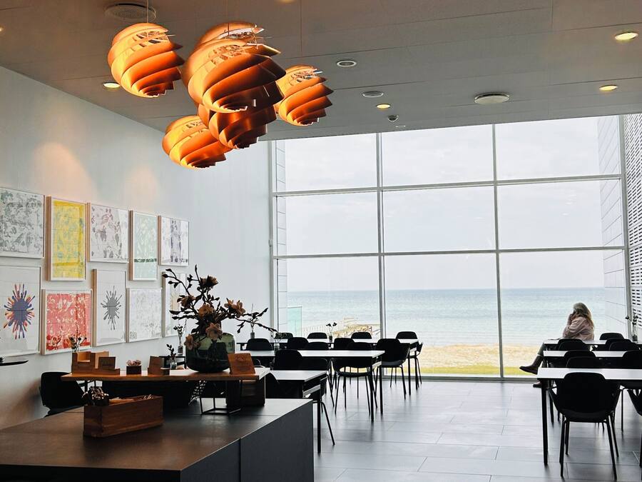 A modern interior with a large window overlooking the sea, a central table with a vase of flowers, and artwork on the walls.