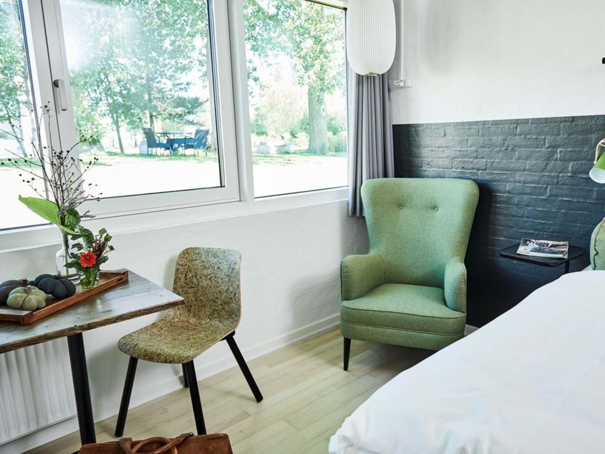 A hotel room with a bed, a green armchair, a small table with a vase of flowers, and a view of the outdoors through large windows.