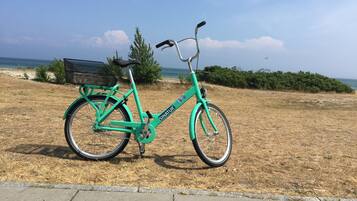 A turquoise bicycle with a basket and a seat, parked on a concrete path near a grassy area with a bench and a tree.
