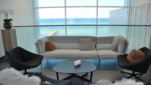 Una sala de estar moderna con un sofá gris, dos sillones negros, una mesa de centro redonda y vistas al océano a través de amplios ventanales.