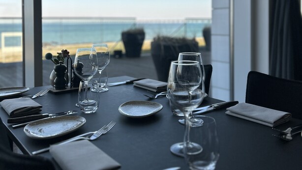 Mesa preparada para comer con vista al mar y un puente al fondo.