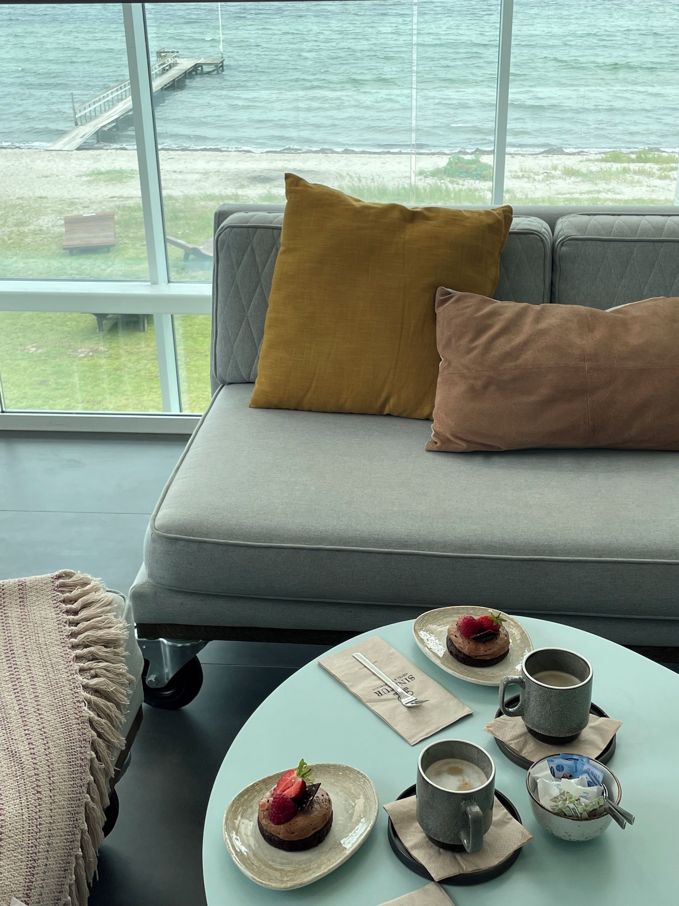 Una habitación con sofá, una mesa con postre, tazas de café y vista al mar.