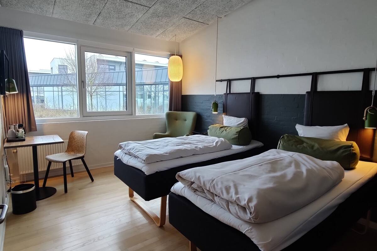 A hotel room with two beds, a desk, a chair, and a view of buildings through large windows.