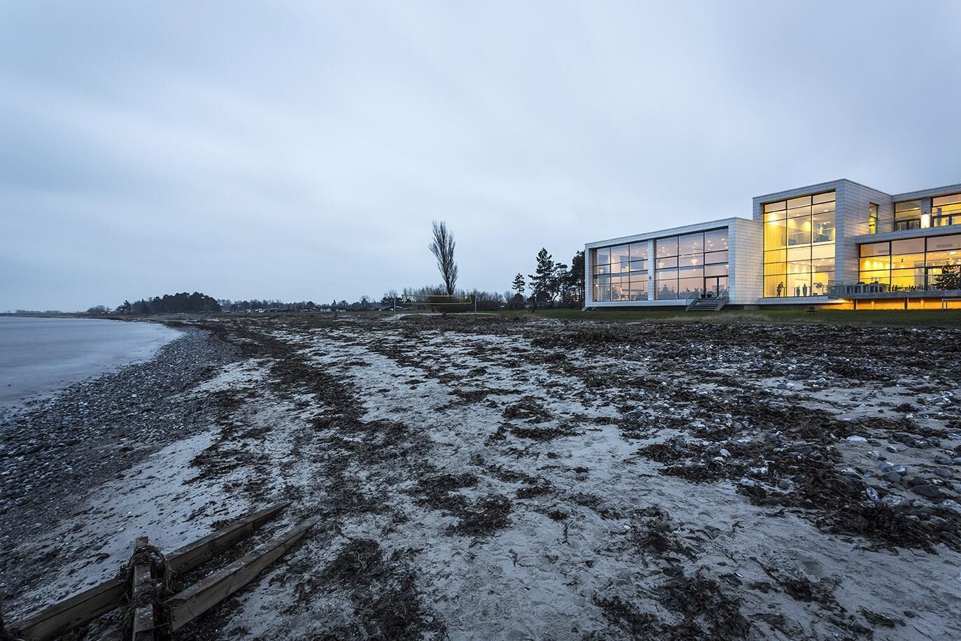 Un edificio moderno con grandes ventanales, una valla de madera y una playa al fondo.