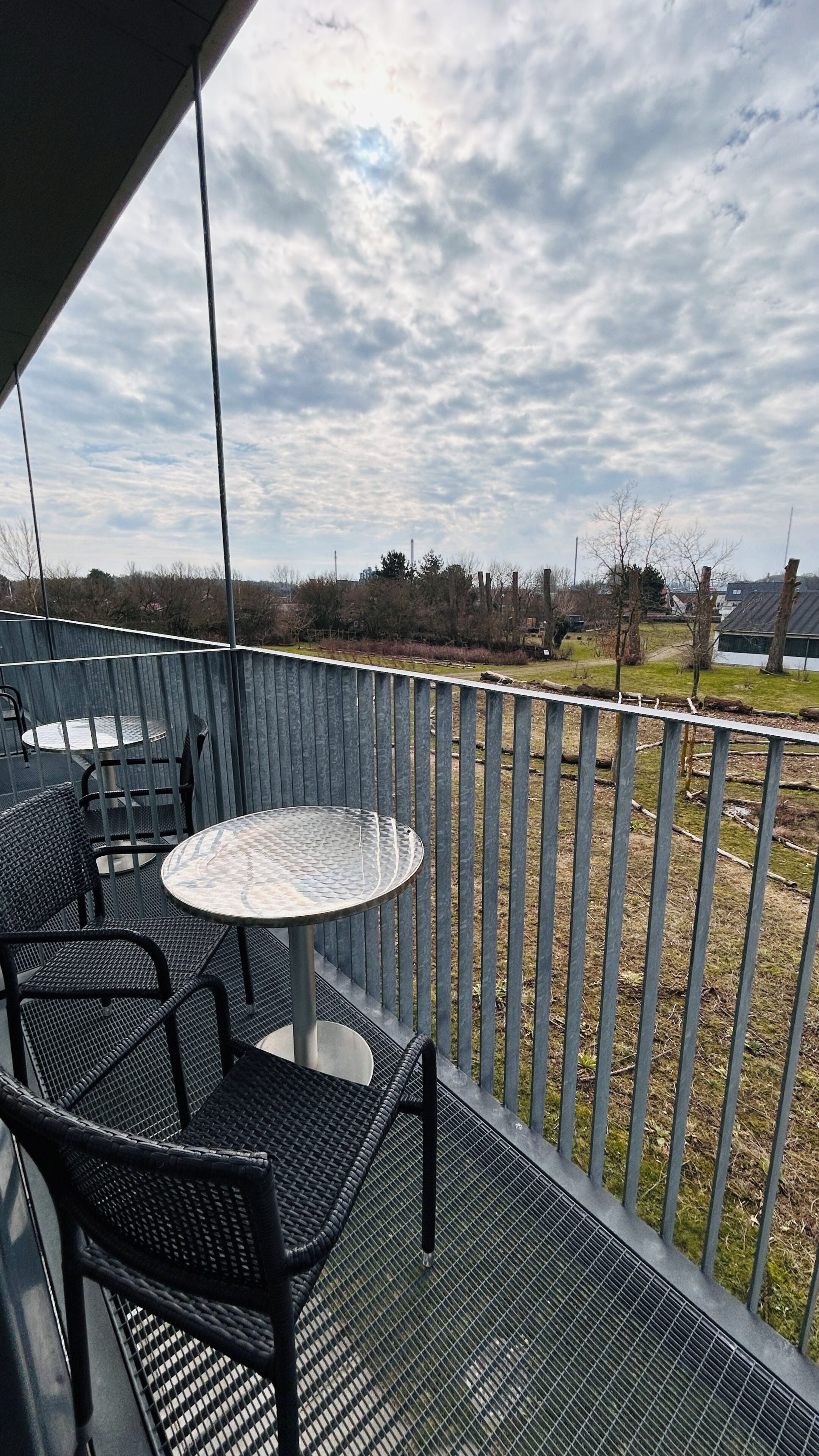 Un balcon avec une table ronde et des chaises, une rambarde en métal, et une vue sur un espace verdoyant avec des arbres et des bâtiments au loin.