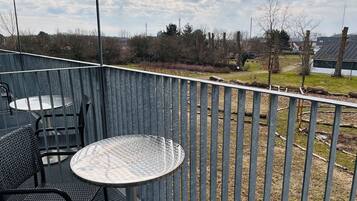 Un balcon avec une table ronde et des chaises, une rambarde en métal, et une vue sur un espace verdoyant avec des arbres et des bâtiments au loin.