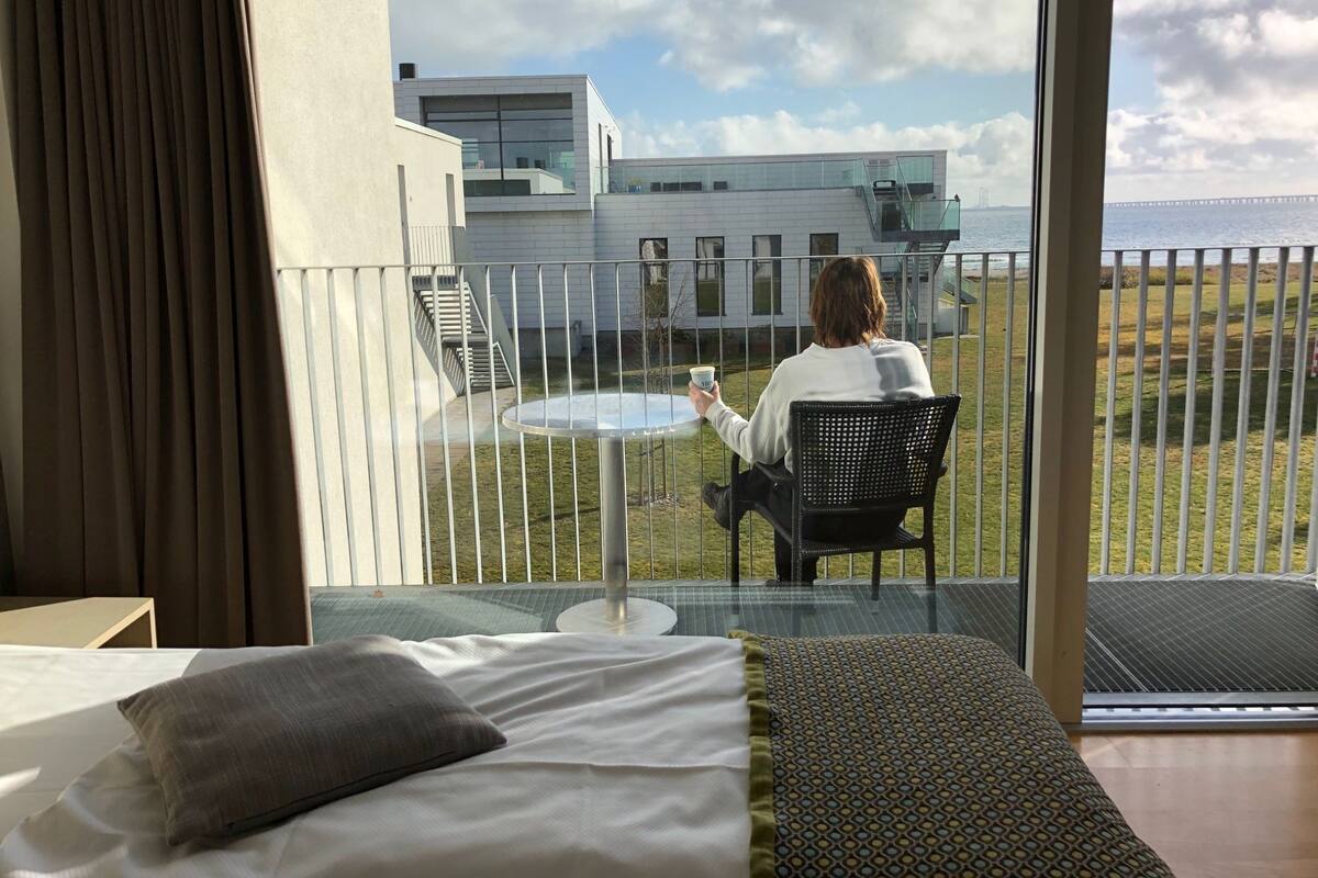 A hotel room with a balcony, a bed, a small table, and a person sitting outside holding a cup.