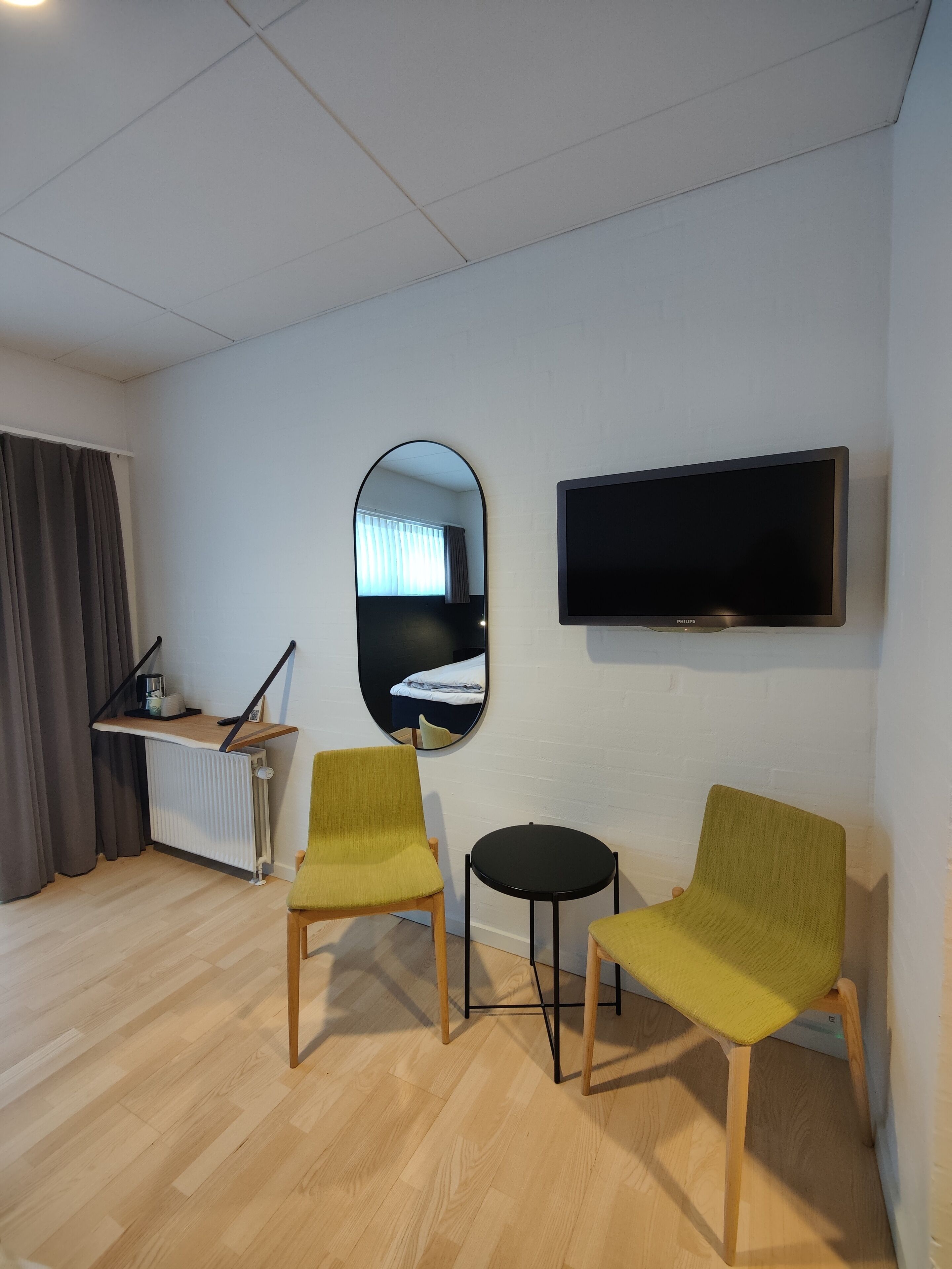 A modern room with two green armchairs, a small black table, a flat-screen TV, and a mirror reflecting a bed and window.
