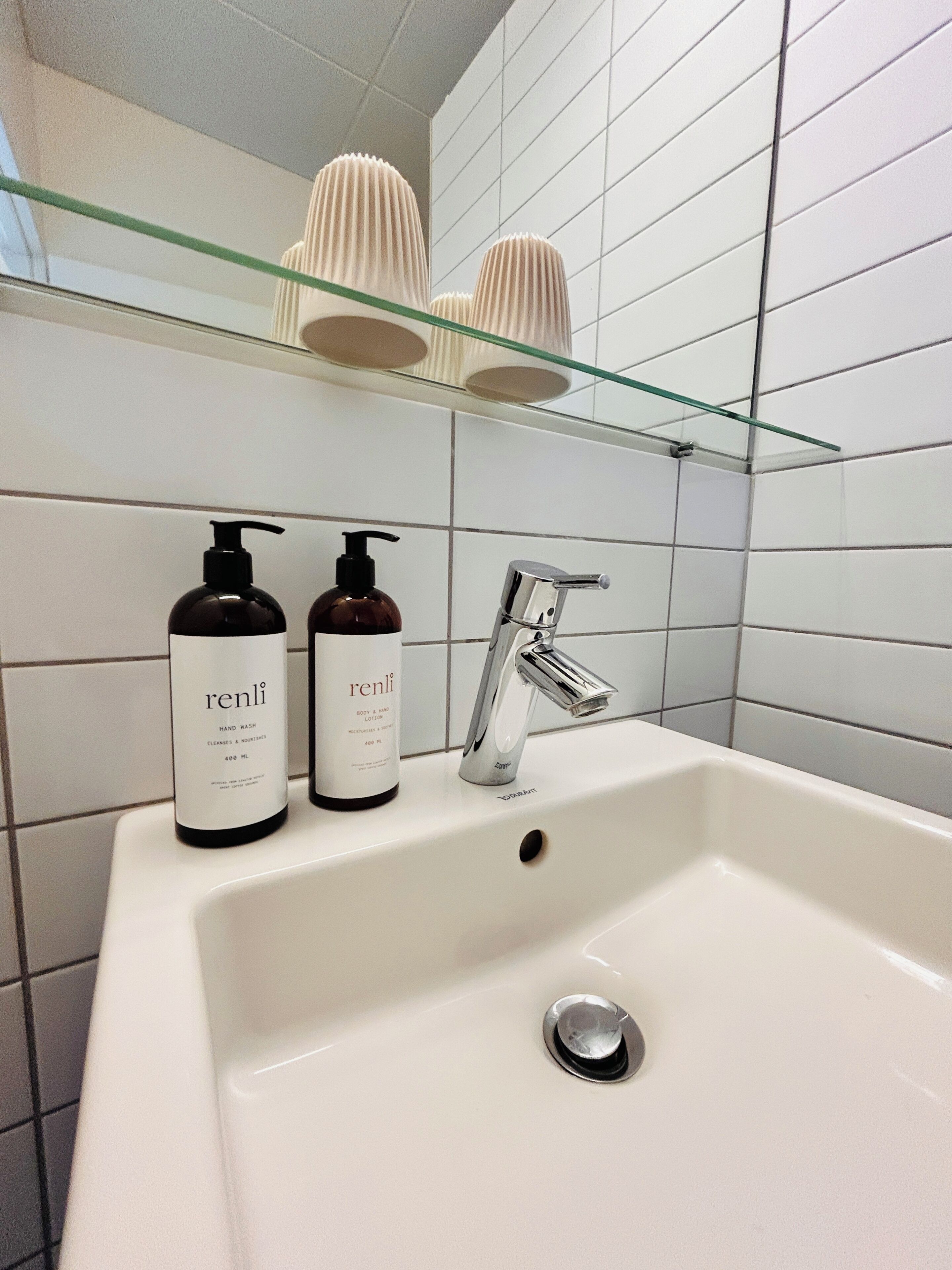 Un baño con un lavamanos blanco, dos botellas de jabón corporal Renli, un grifo cromado y un estante de vidrio con objetos decorativos.