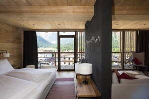 A wooden room with a bed, a sofa, a table, and a view of mountains. - Berghotel (Sesto)