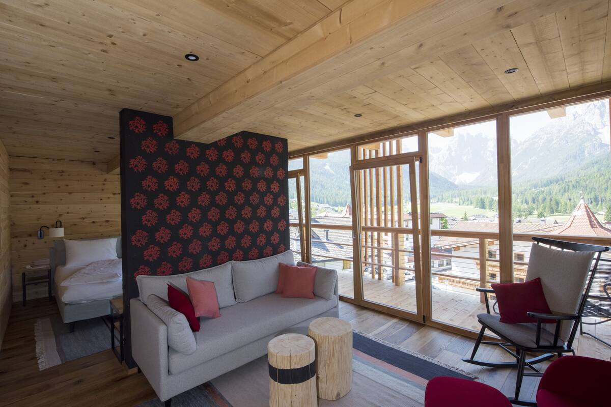 A cozy cabin interior with a wooden ceiling, a sofa, a rocking chair, and a large window offering a mountain view.