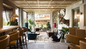 A spacious indoor area with a stone wall, wooden ceiling, and a mounted deer head. There are multiple seating areas with chairs and tables, and a bar counter with stools. - Berghotel (Sesto)