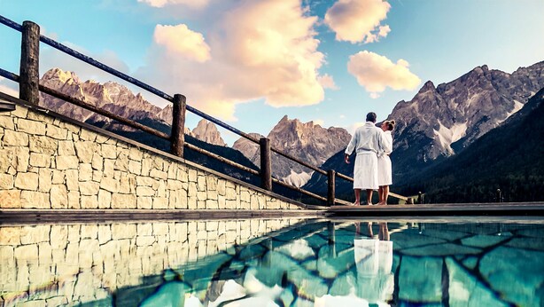 A stone wall with a wooden railing overlooking a clear pool reflecting mountains. - Berghotel (Sesto)