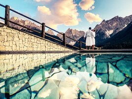 Un muro de piedra con barandilla de madera que da a una piscina cristalina que refleja montañas.
