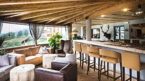 A spacious living area with a wooden ceiling, a sofa, a coffee table, and a bar with stools. - Berghotel (Sesto)
