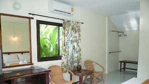 A hotel room with a wooden desk, two wicker chairs, a bed with a white towel, a large mirror, a window with floral curtains, and an air conditioning unit.