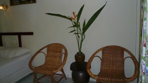 A room with two wicker chairs, a bed, a potted plant, and a painting on the wall.