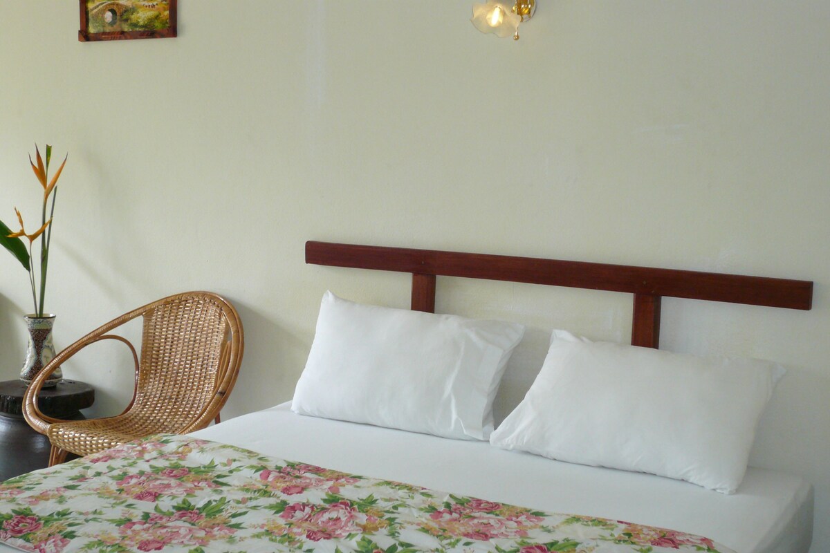 A neatly made bed with white bedding and a floral-patterned duvet cover, a wooden headboard, a wicker chair, and a small table with a vase of flowers.