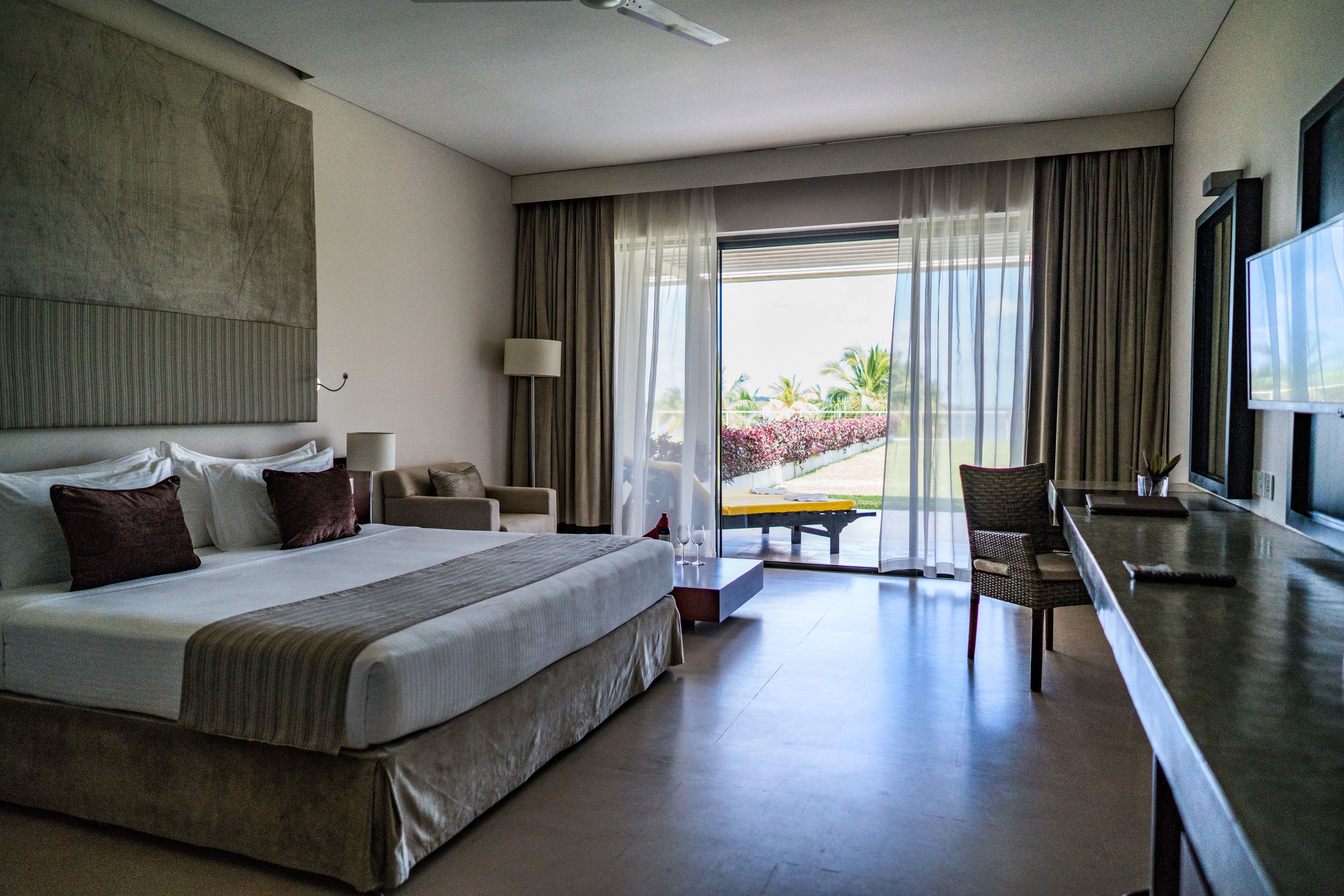 A modern hotel room with a large bed, a flat-screen TV, a seating area with a table and chairs, and a view of the outdoors through large windows.