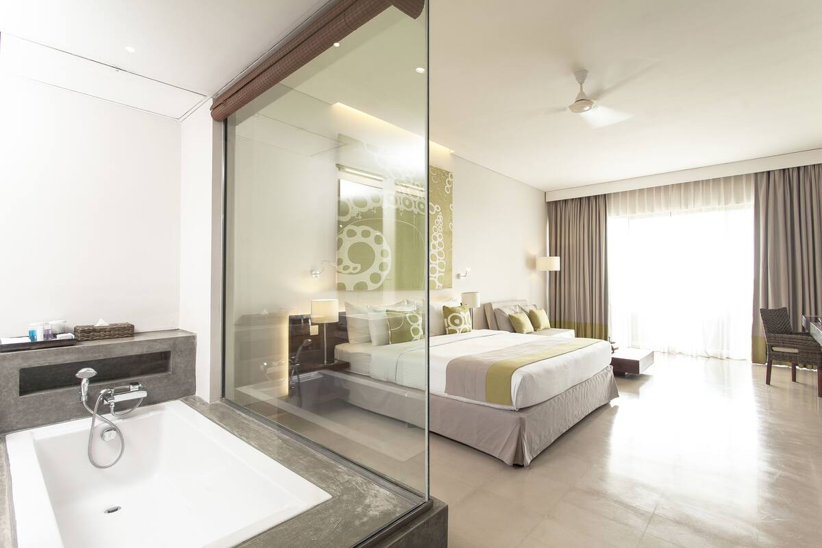 A modern hotel room with a large bed, a glass-enclosed shower, a large window with curtains, and a seating area.