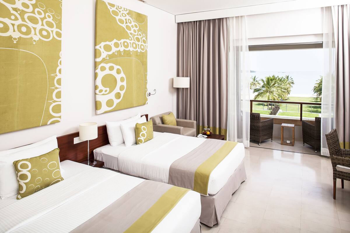 A hotel room with two beds, a sofa, a balcony with a view of palm trees, and a large abstract painting on the wall.