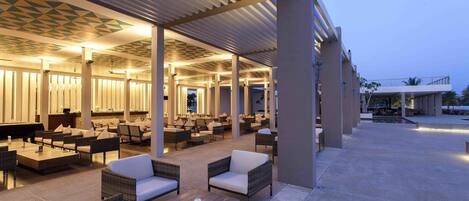 A spacious outdoor lounge area with wicker furniture and a covered seating area, featuring a patterned ceiling and ambient lighting.