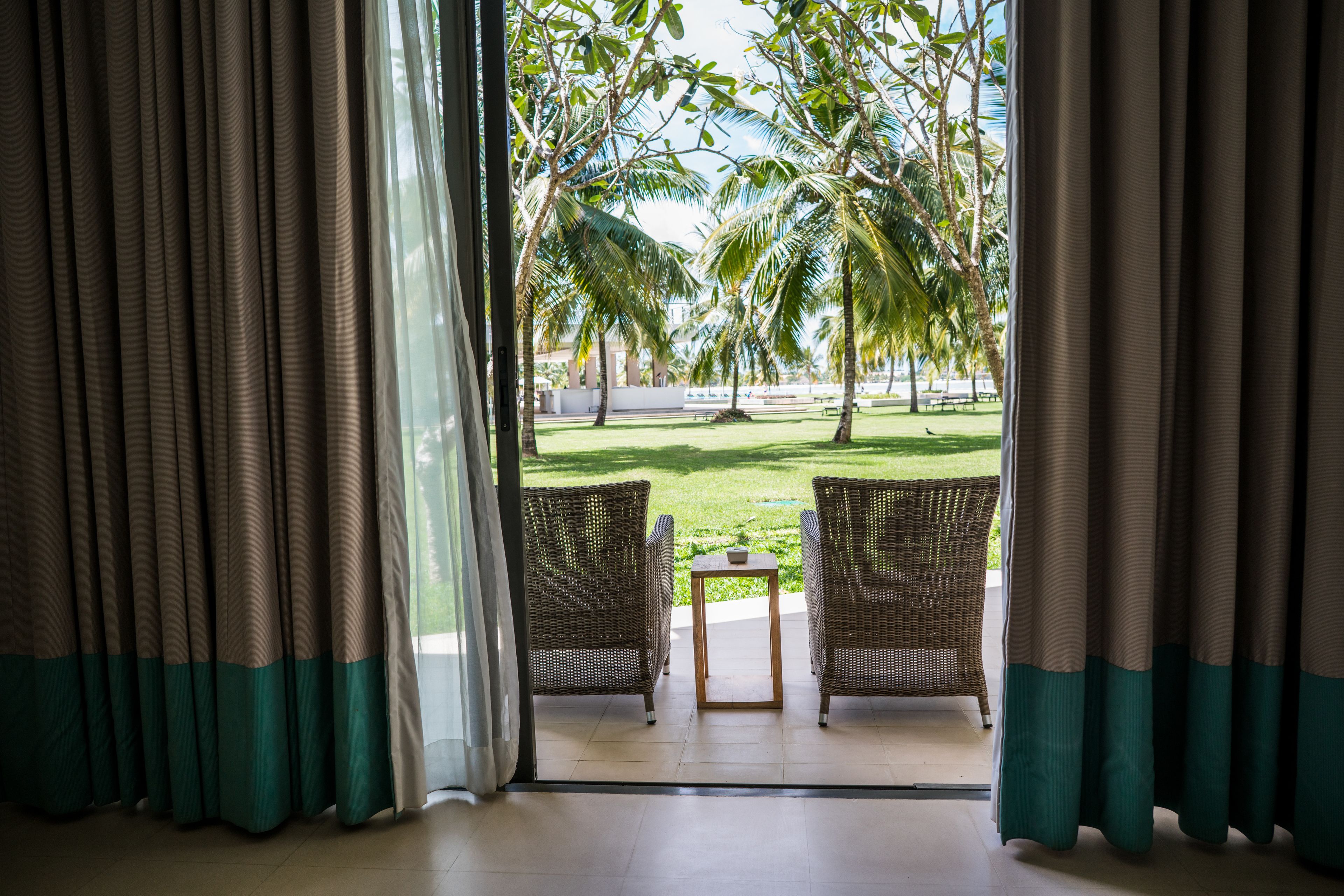 A view from a room with two wicker chairs and a small table, looking out to a grassy area with palm trees and a white building in the distance.
