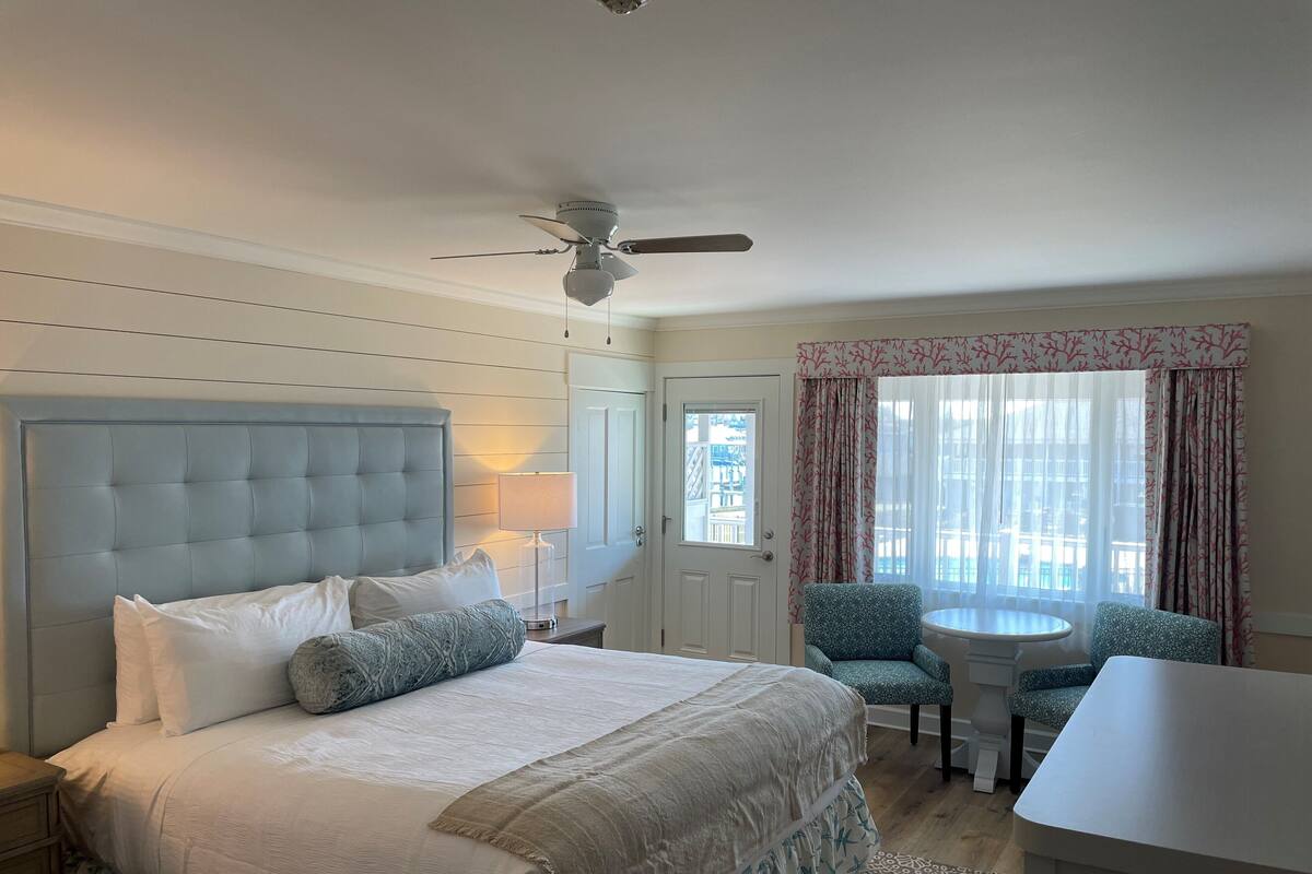 Una habitación de hotel con una cama grande, un ventilador de techo, una mesita y una silla.