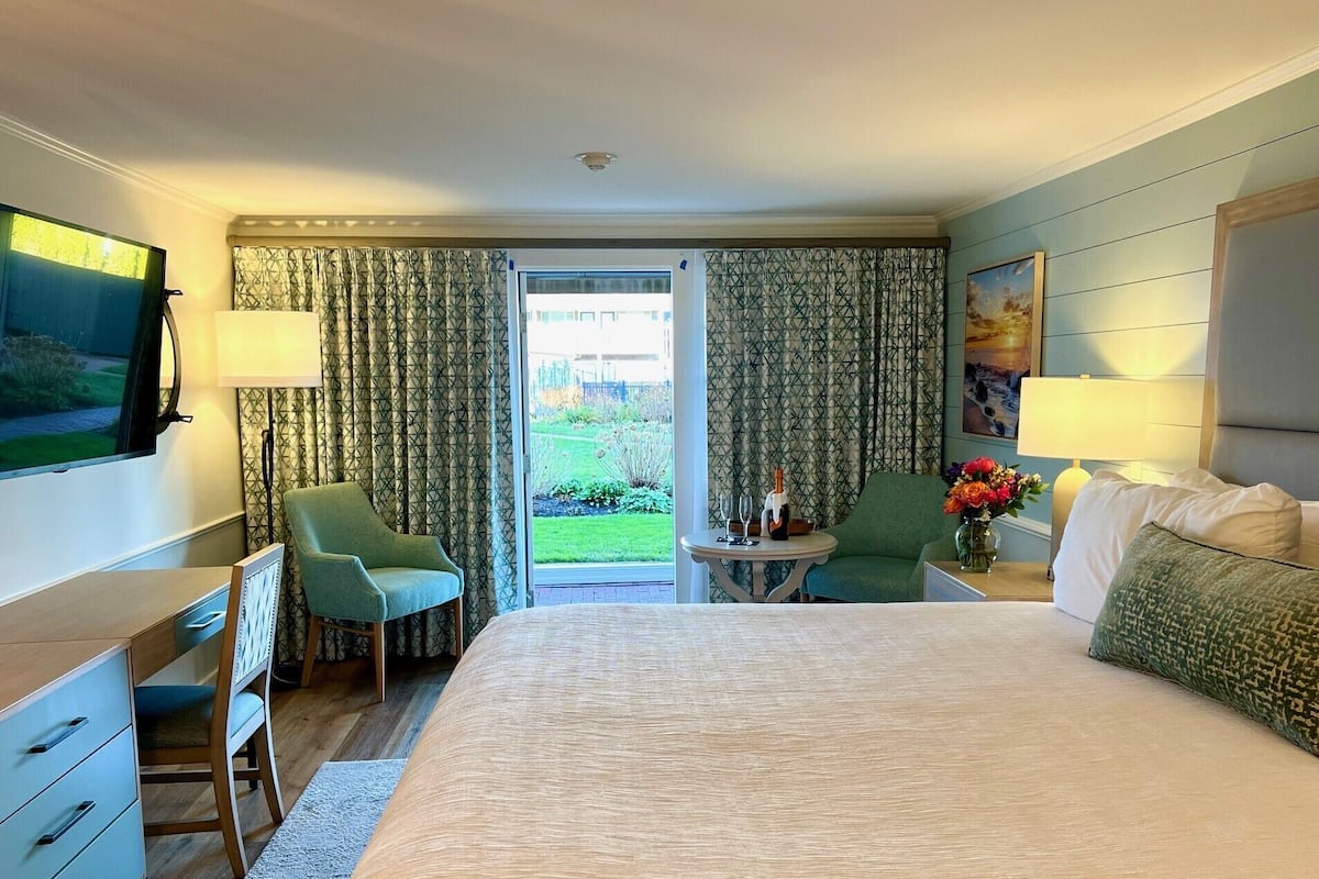 Habitación de hotel con una cama grande, un escritorio, una silla, una mesita con una botella y un vaso, y vistas a un jardín a través de la puerta.